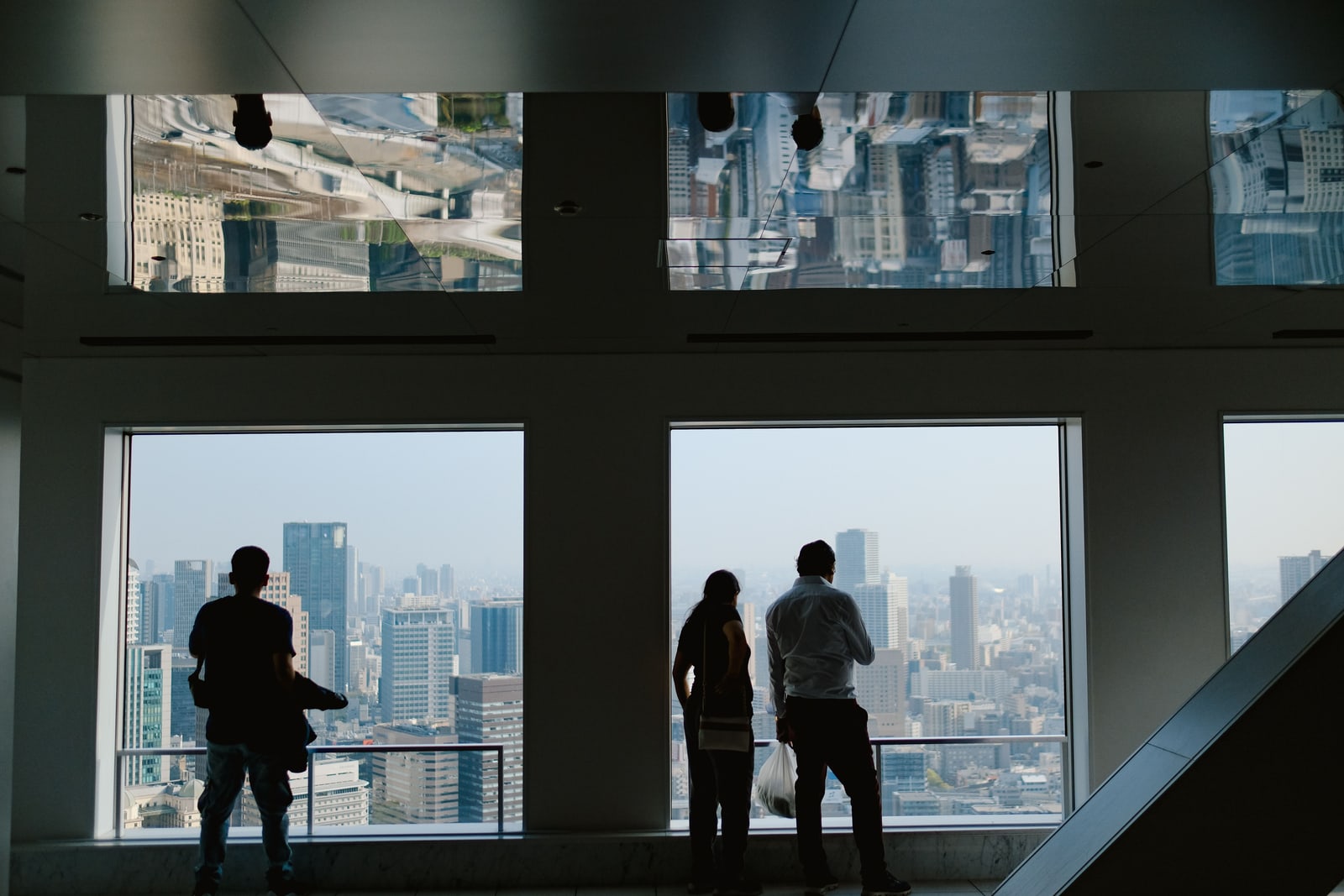 Three visitors in silhouette standing at floor-to-ceiling windows of an indoor high-rise viewing floor, the city below.