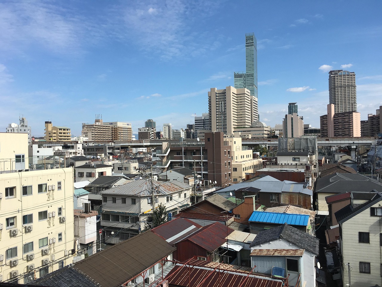 Low tiled and metal rooftops of a residential Osaka block, with Abeno Harukas rising high above them in the distance under a blue sky.