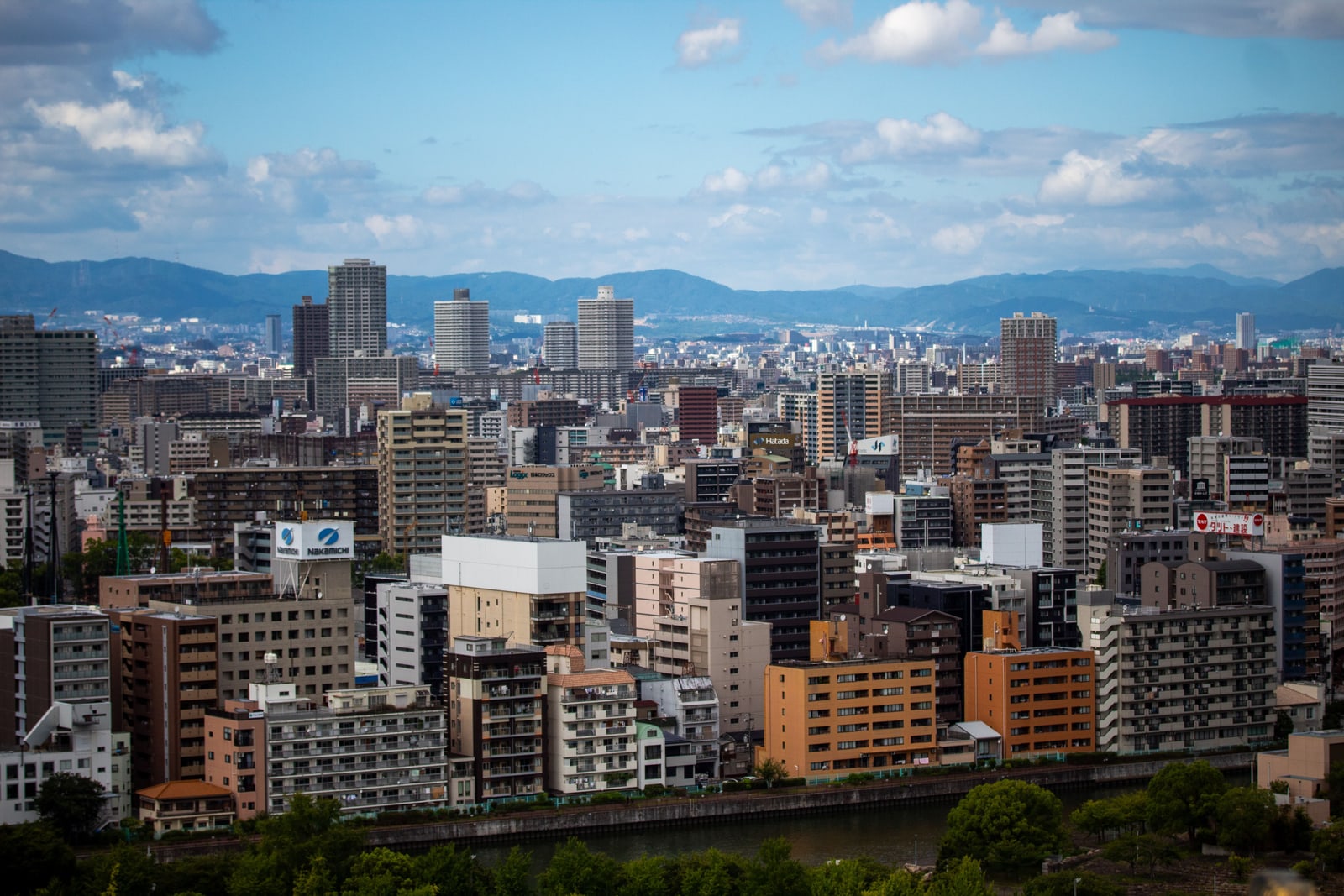 Wide daytime view over Osaka apartment and office blocks to a mountain range under broken cloud.