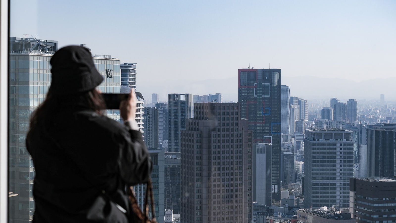A visitor in a hat, seen from the side with the face hidden behind a phone, photographing Osaka towers through a high window.