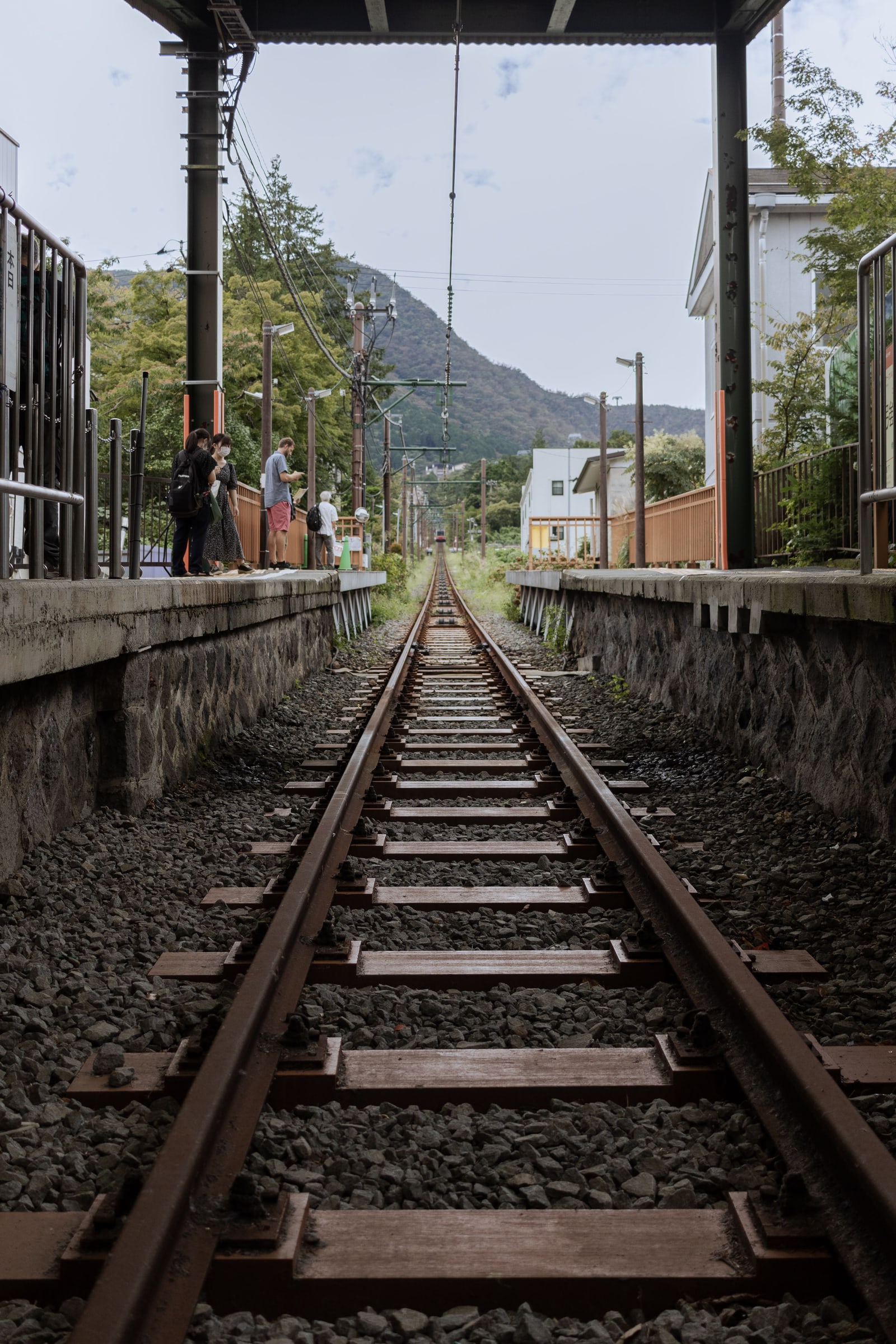 A single railway track running between two small platforms toward wooded hills in Hakone, with two small figures at the far end.