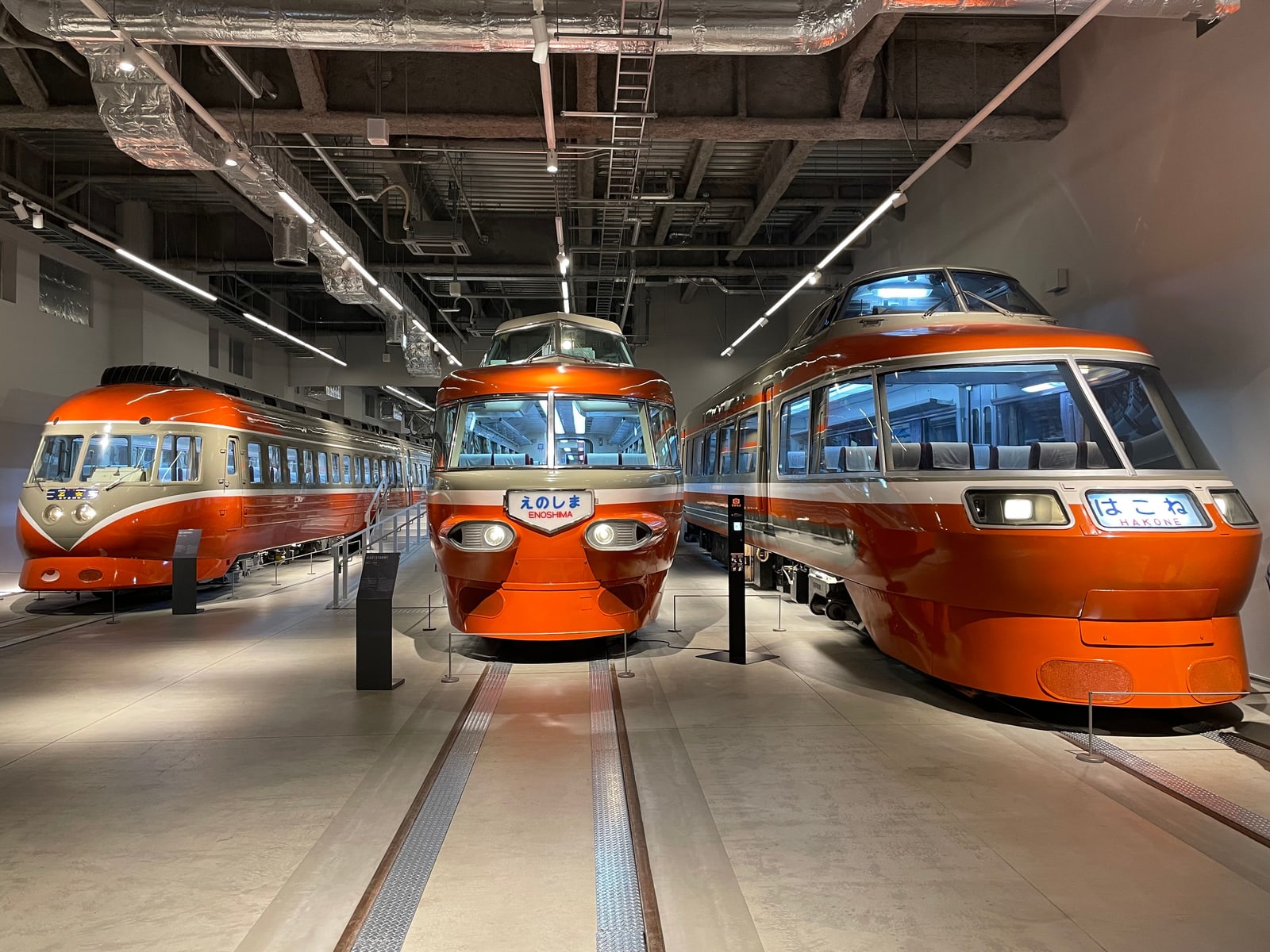 Three retired orange-and-white Romancecar trains on display indoors, their rounded front cabs facing the camera, with destination boards reading はこね and えのしま.