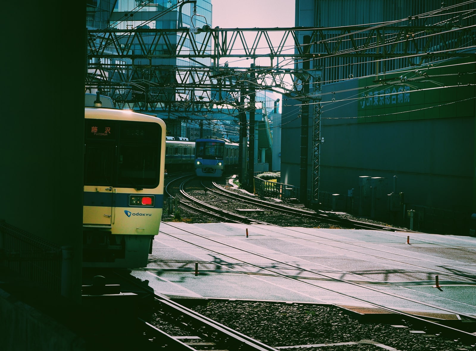 The front of a cream-colored Odakyu commuter train with a blue stripe and the odakyu logo, under station wires between tall buildings.