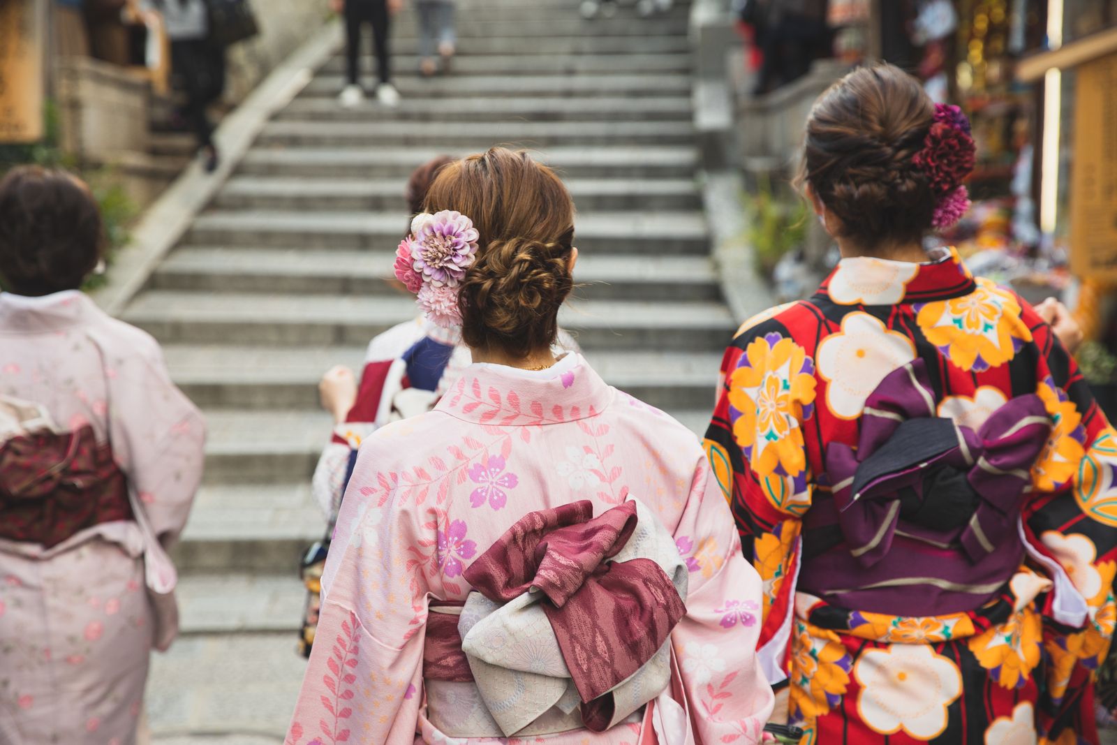 Three women in kimono seen from behind at the foot of a broad stone staircase, two of them with dressed updos and fabric flower ornaments, the center figure's obi tied in a wine-colored bow.