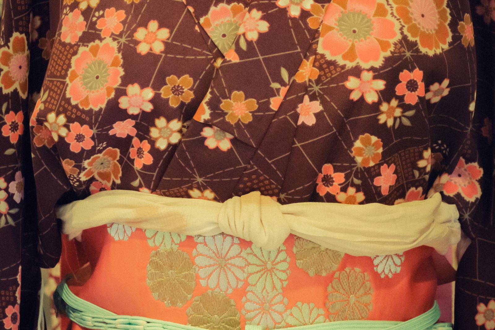 A close view of a kimono at the waist: a dark floral outer layer, a coral obi woven with gold roundels, and a cream cloth knotted at the center.