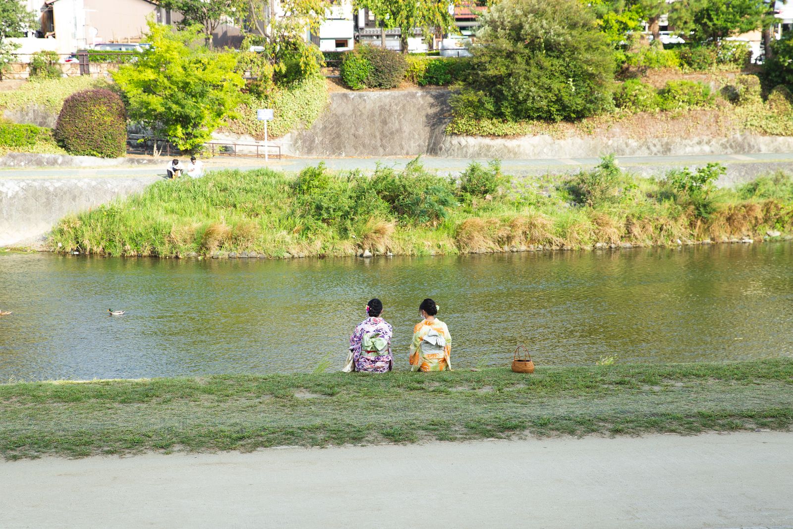 Two women in kimono sitting side by side on a grass riverbank with their backs to the camera, a woven basket bag on the grass beside them, a stepped stone embankment on the far bank.