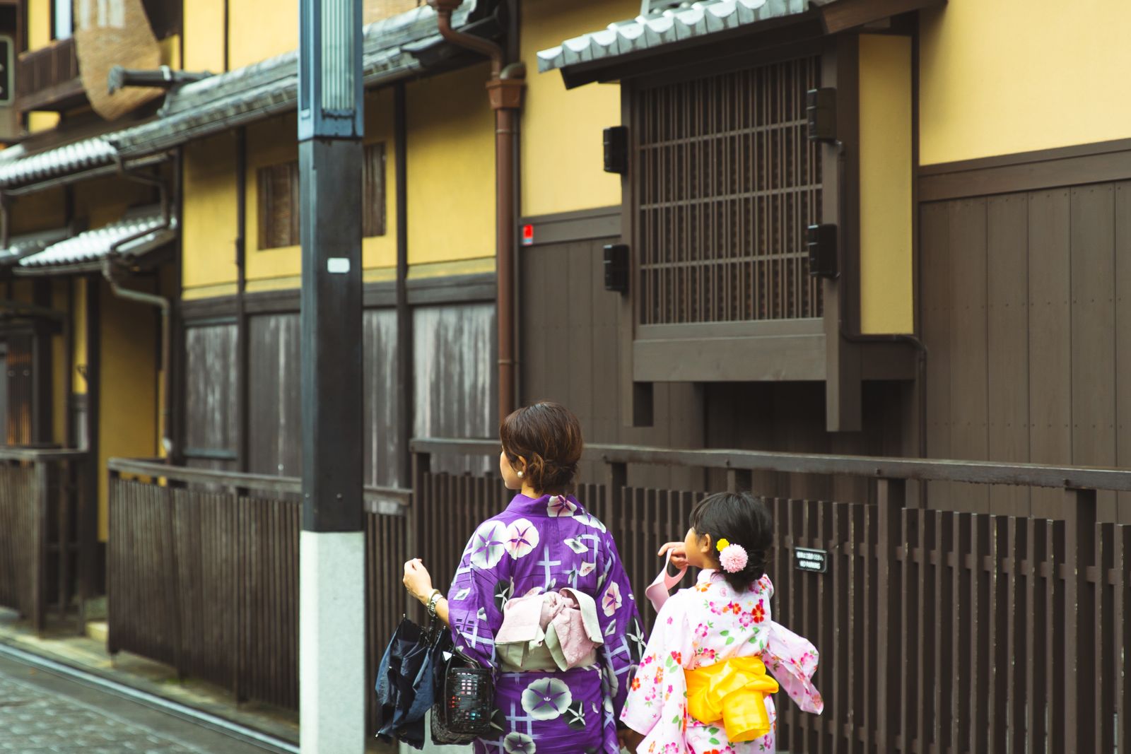 Two people in Japanese dress walking away along a quiet street of ochre-plastered machiya with dark timber lattice.