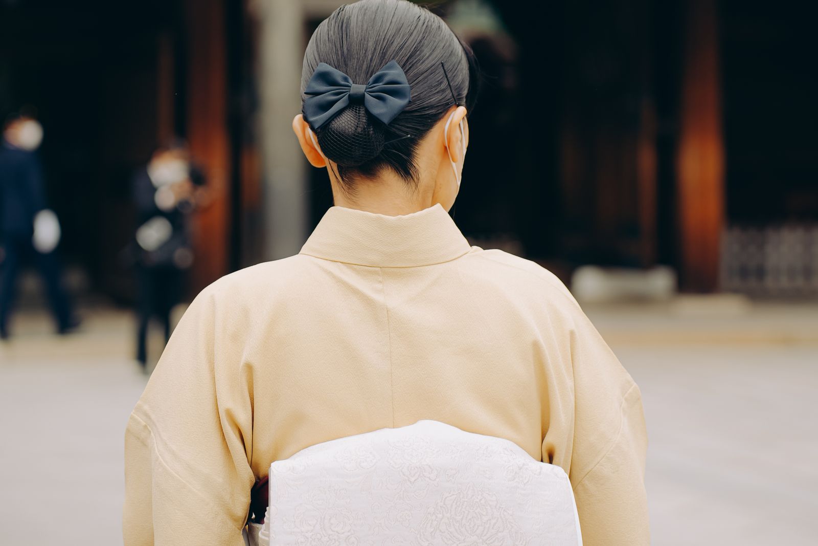 A close view from behind of a woman in a plain cream kimono with a white brocade obi, her hair gathered into a low chignon held by a navy fabric bow, the background thrown fully out of focus.