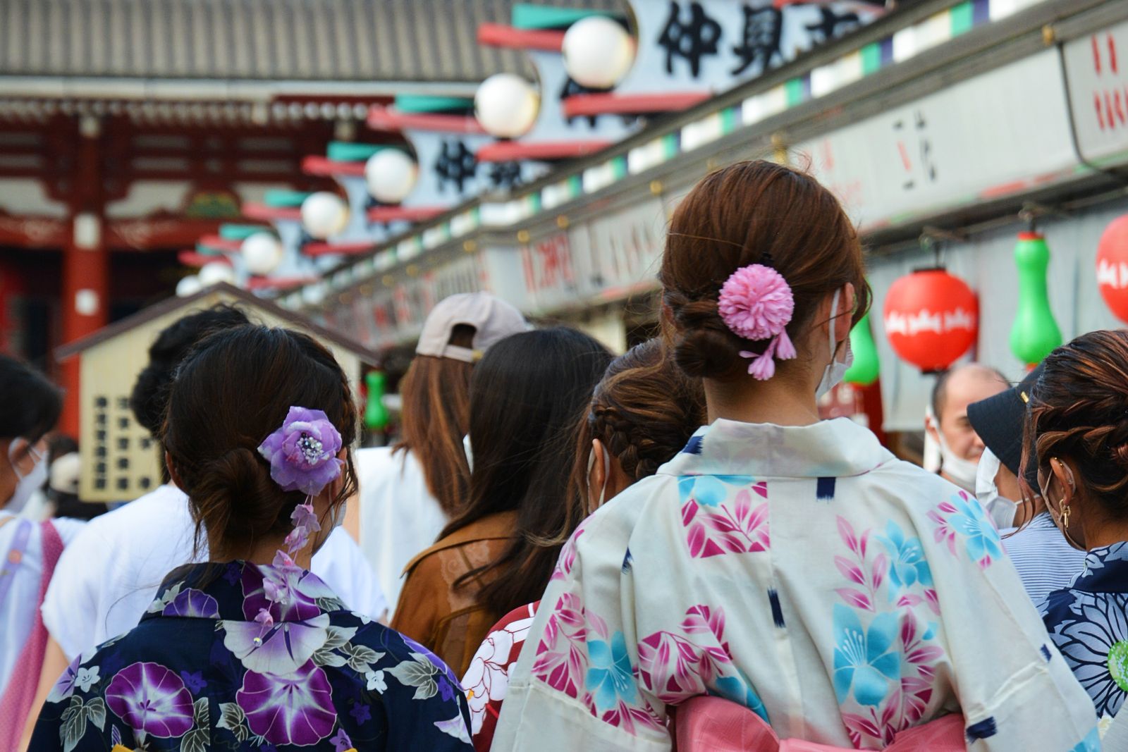 A crowded covered shopping approach with painted eaves, paper lamps and red lanterns overhead, several women in yukata standing with their backs to the camera, and a large sign reading Nakamise.