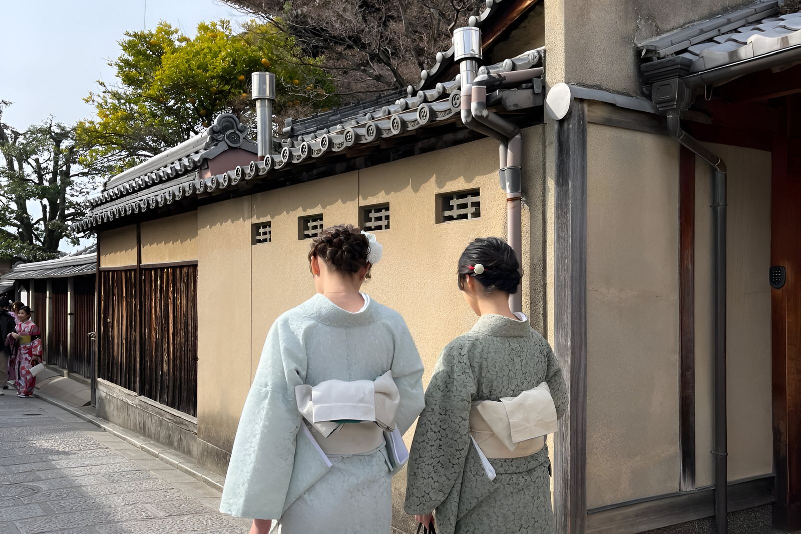 Two women in pale mint and sage kimono with wide cream obi walking away along a stone-paved street beside a long ochre plastered wall topped with a gray tiled roof.