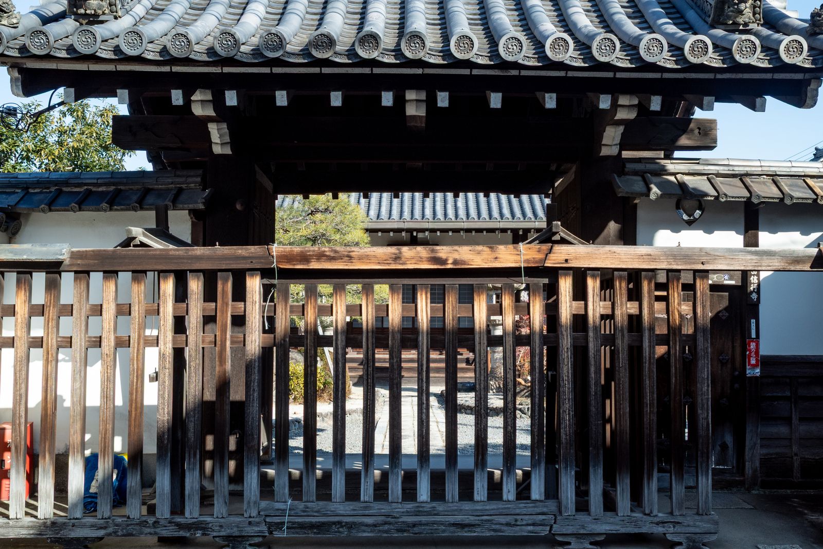 A temple gate seen square-on, with a heavy tiled roof on dark timber brackets and a closed slatted wooden barrier across the opening, raked gravel visible through the slats.