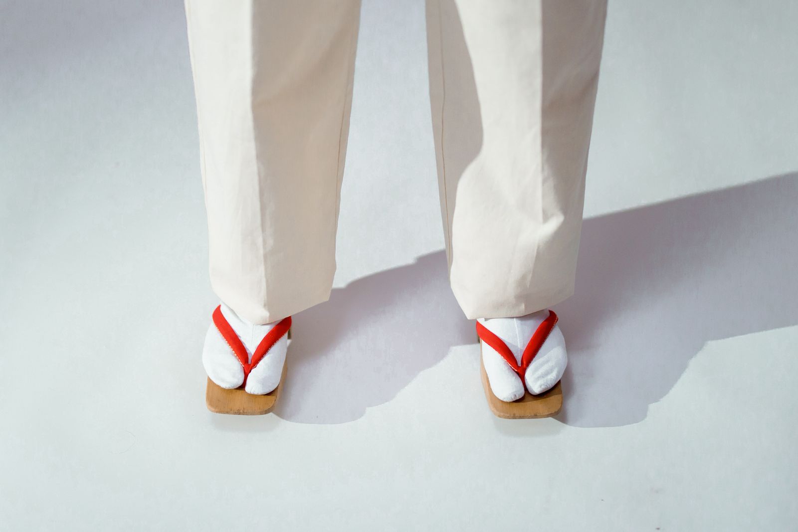 A pair of feet in white split-toe tabi socks standing in wooden thonged sandals with red straps, on a plain pale studio backdrop.