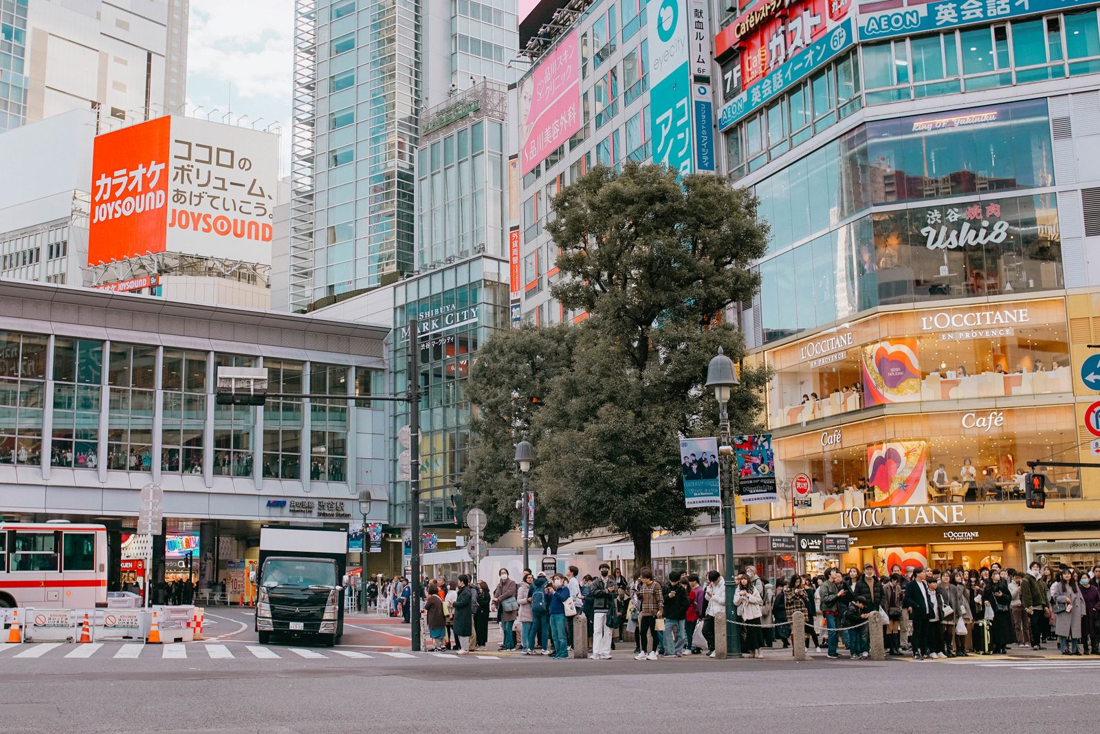 A crowd waiting at a red signal at Shibuya, nobody stepping off the curb, Shibuya Station behind them.