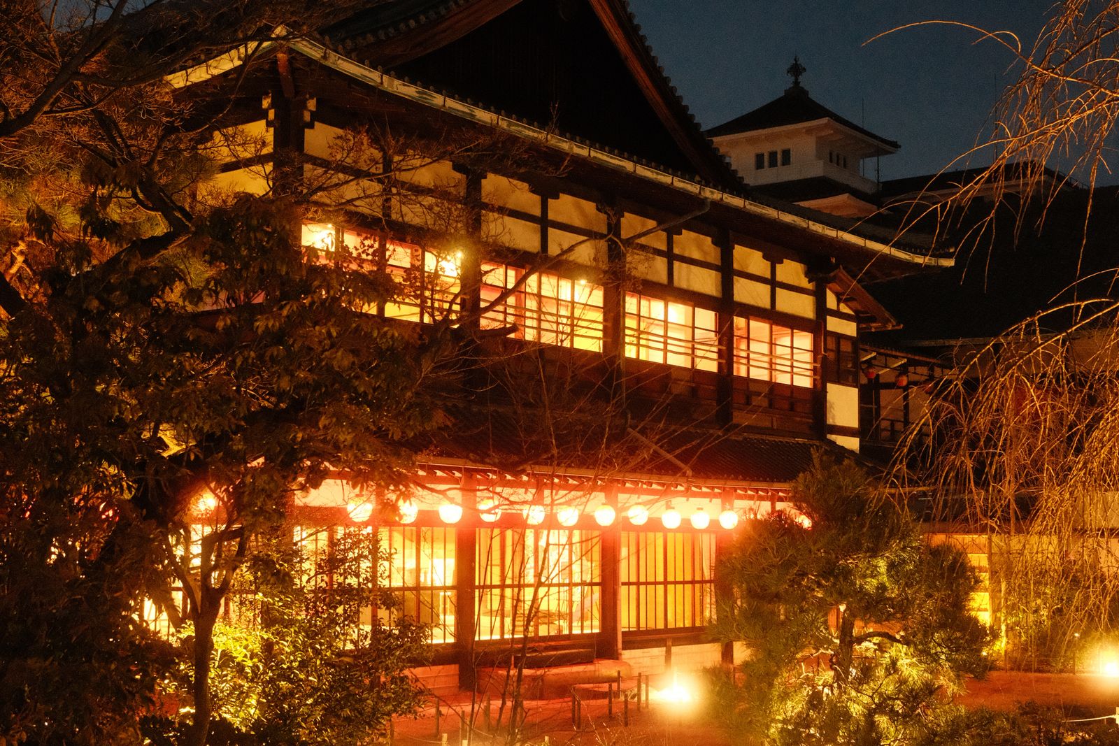 A two-story timber building lit warmly at night, a row of round lanterns along its front and pines beside it.