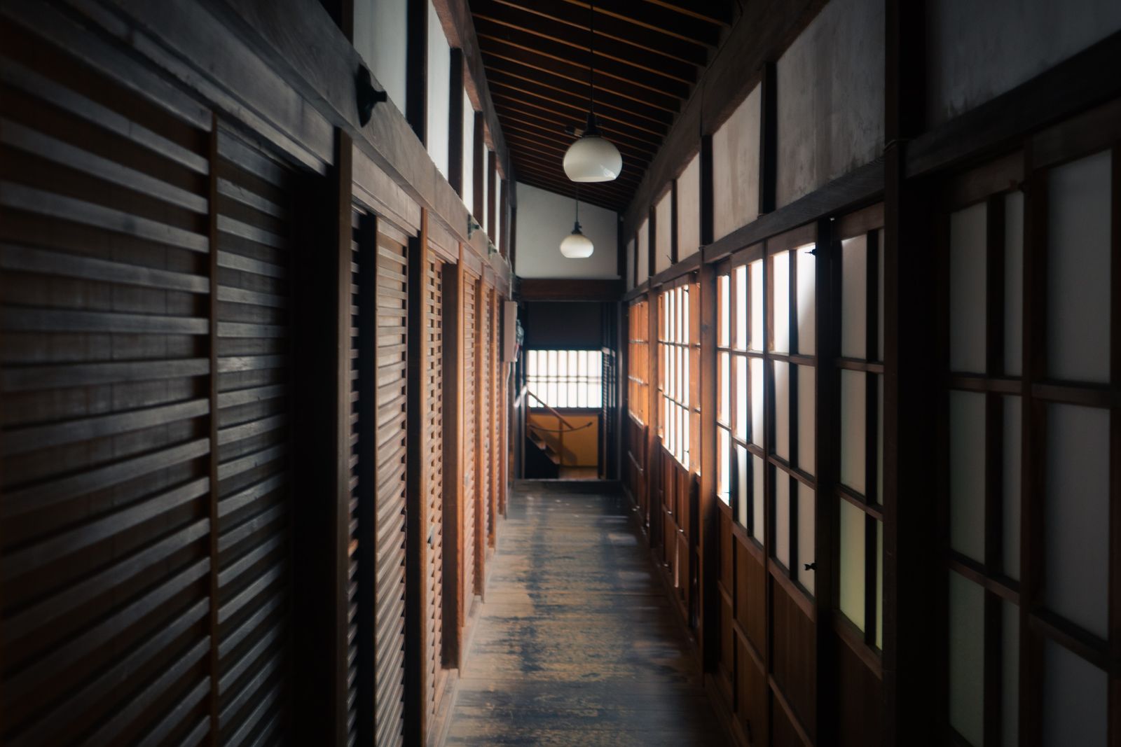 A dark wooden corridor with shoji along one side and two pendant lamps lit overhead.