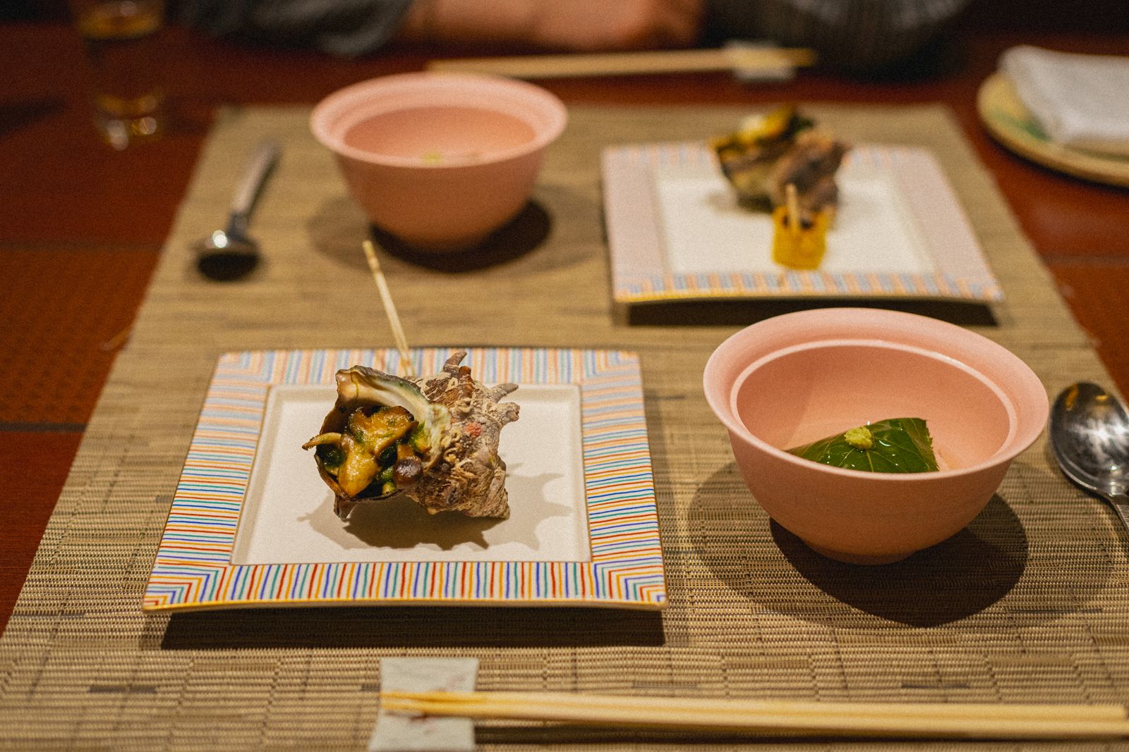 Two place settings on woven mats with a turban-shell dish, lidded bowls and chopsticks.