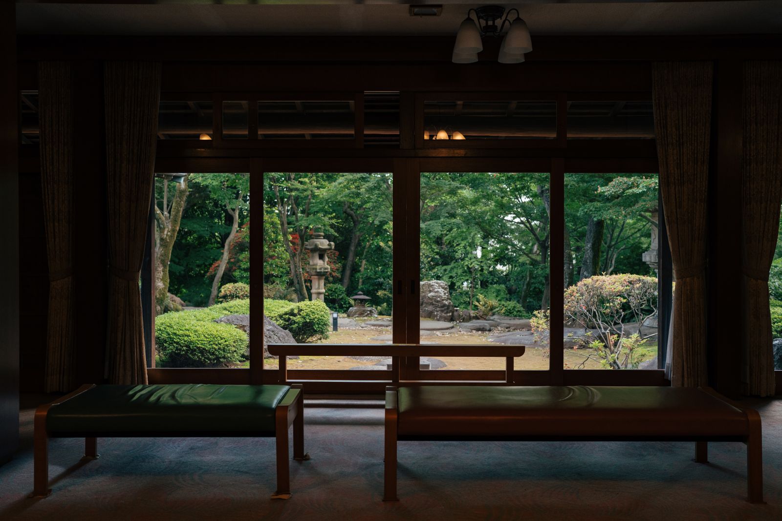 A dim lounge with two leather benches facing floor-to-ceiling windows onto a moss garden with a stone lantern.