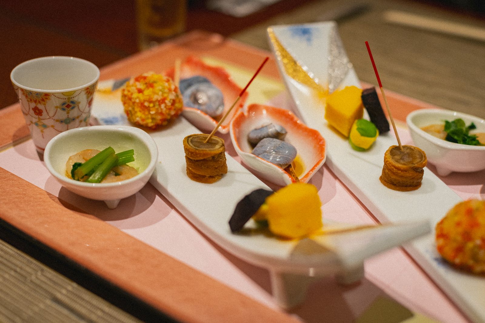 A tray of small appetizer dishes on a pink cloth: skewers, a small bowl and a lidded cup.