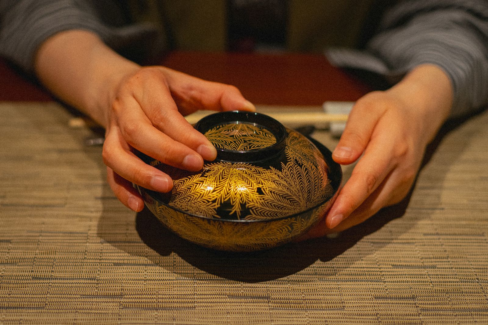 Two hands held around a lidded lacquer bowl, gold pattern on black, resting on a woven mat.