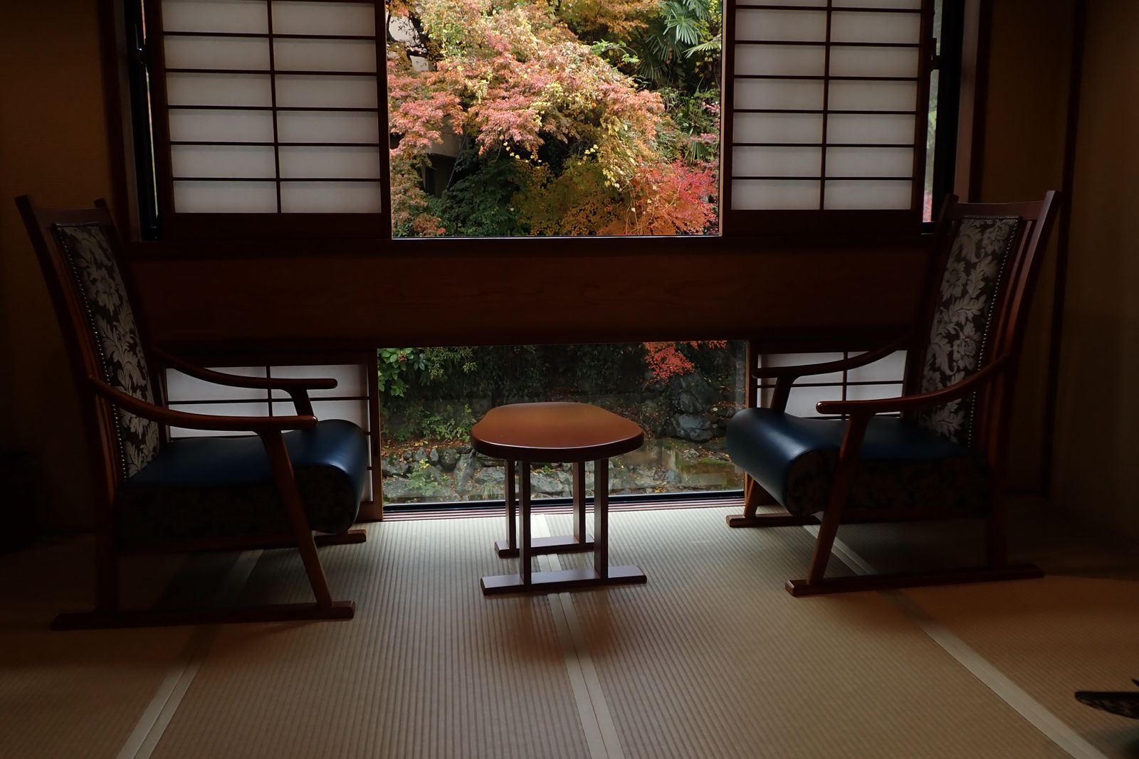 Two upholstered armchairs and a small table in a window alcove, tatami underfoot, autumn maples in the garden beyond.