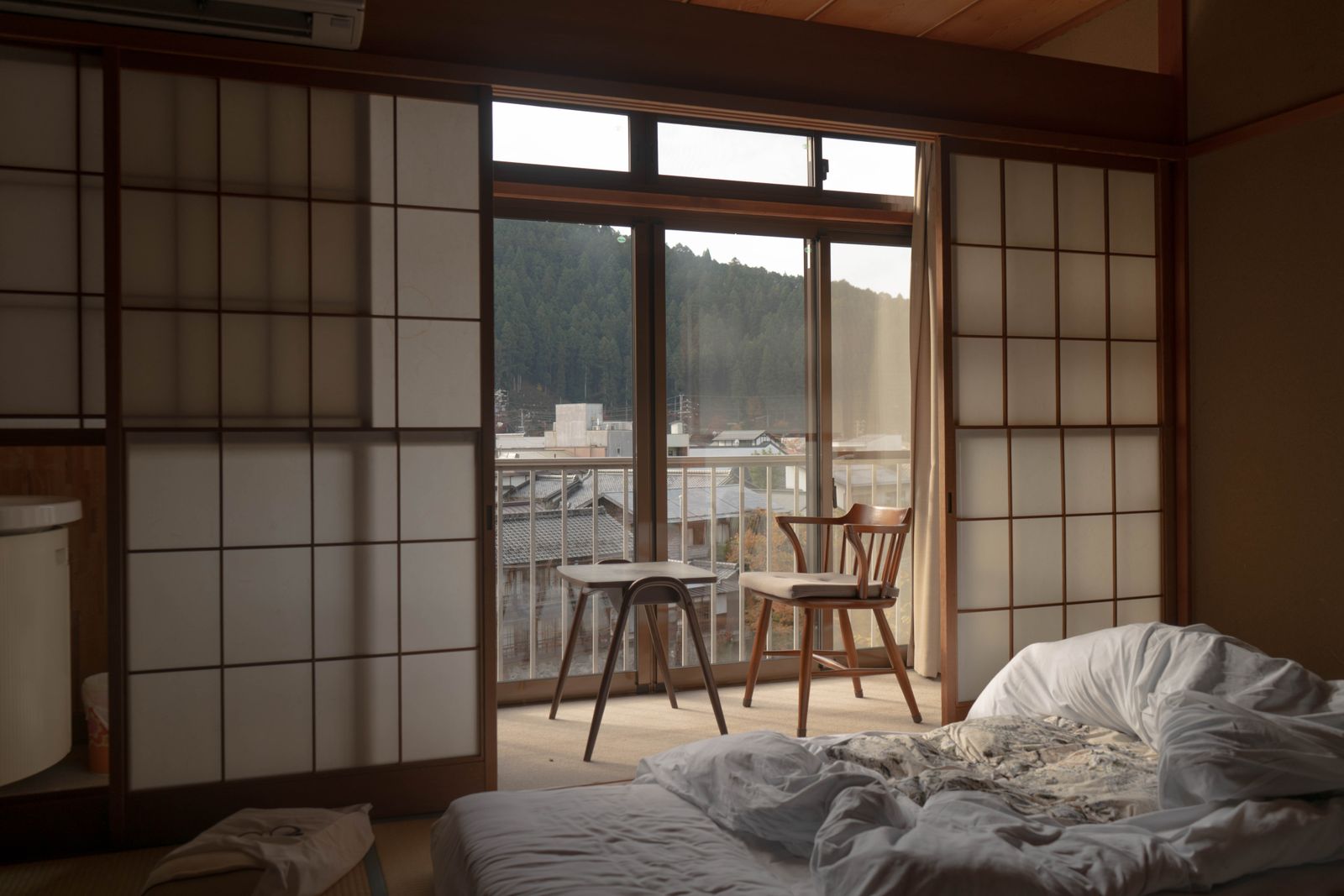 A futon laid out on tatami beside shoji, a window alcove with two chairs, hills outside.