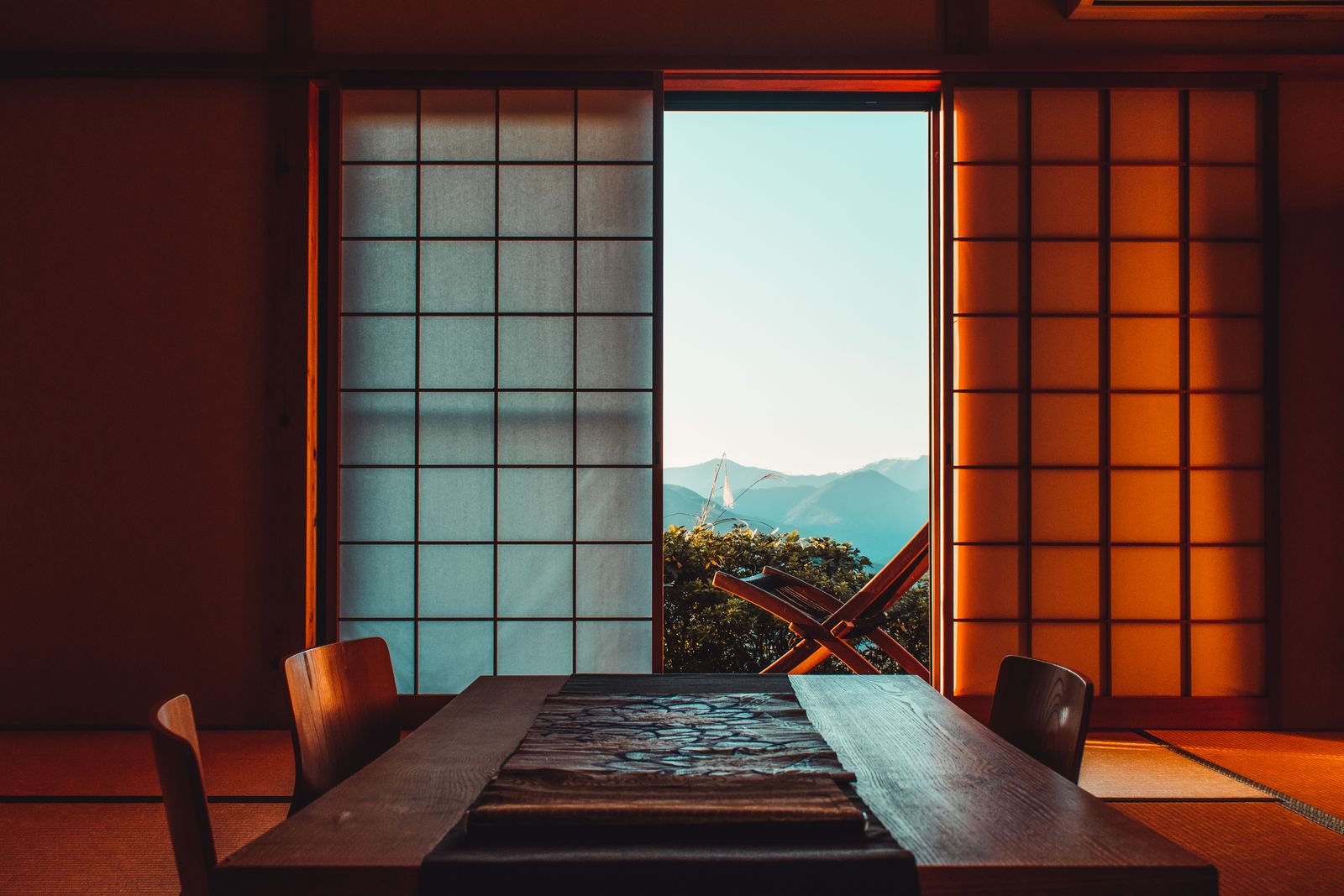A dark wooden table with two chairs before open shoji, mountains beyond in golden-hour light, a folding chair on the veranda.