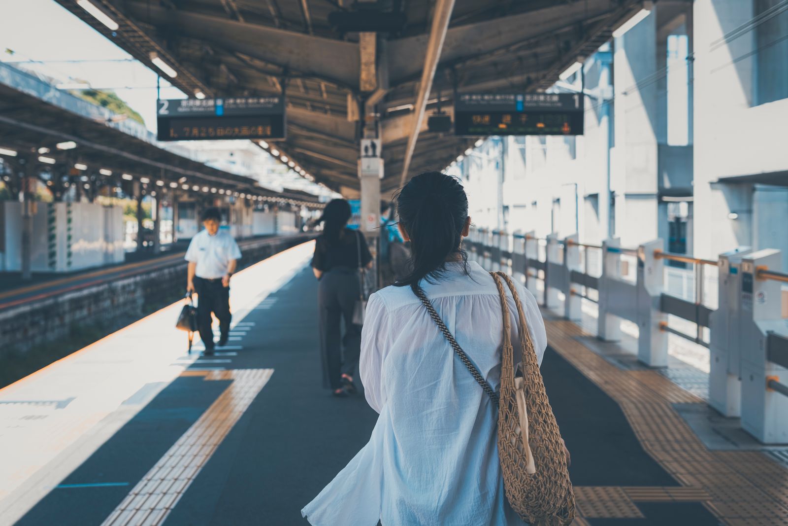 A Tokyo station platform in daylight, commuters seen from behind, line signage overhead.