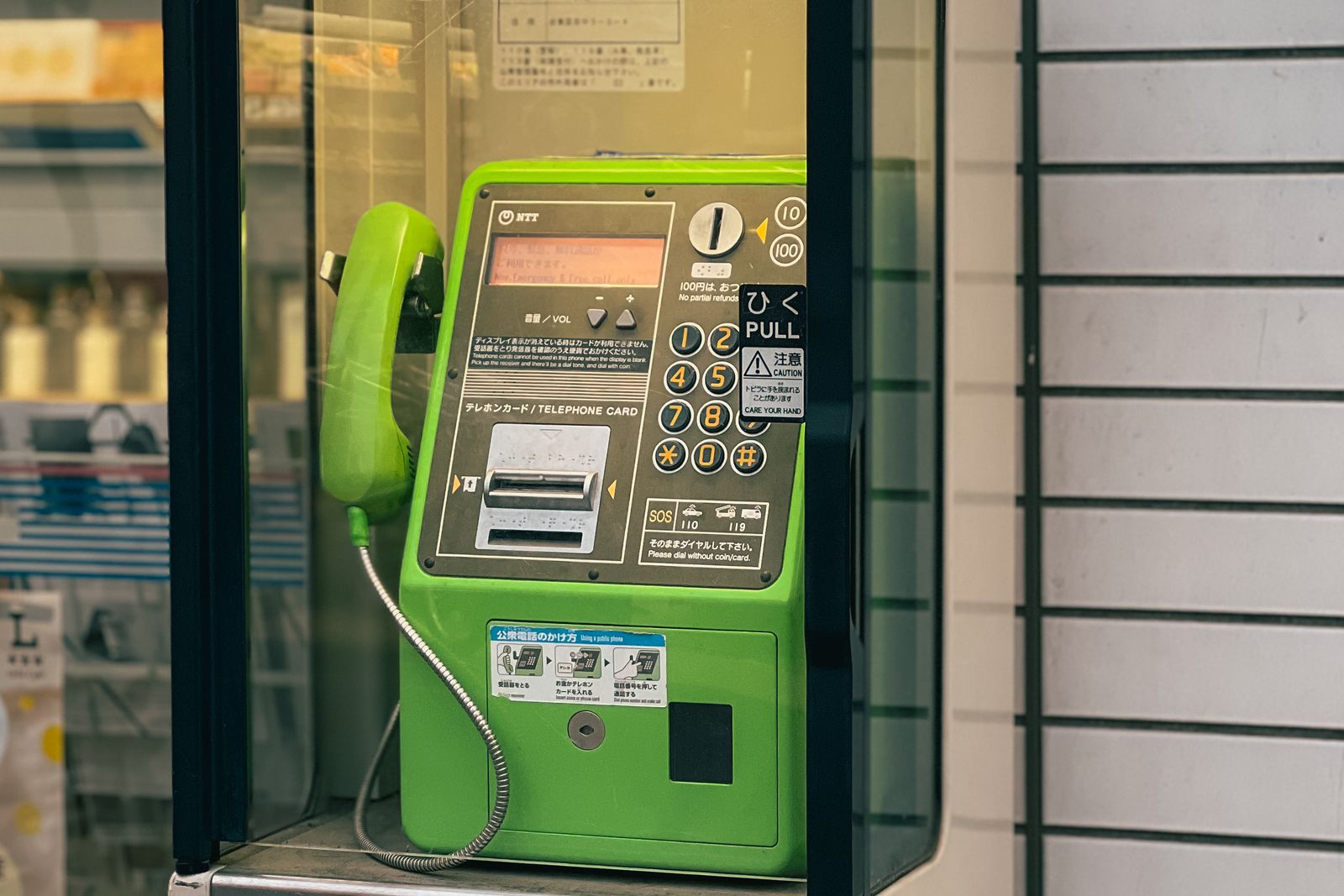 A green NTT public payphone on a street, its emergency panel showing 110 and 119.