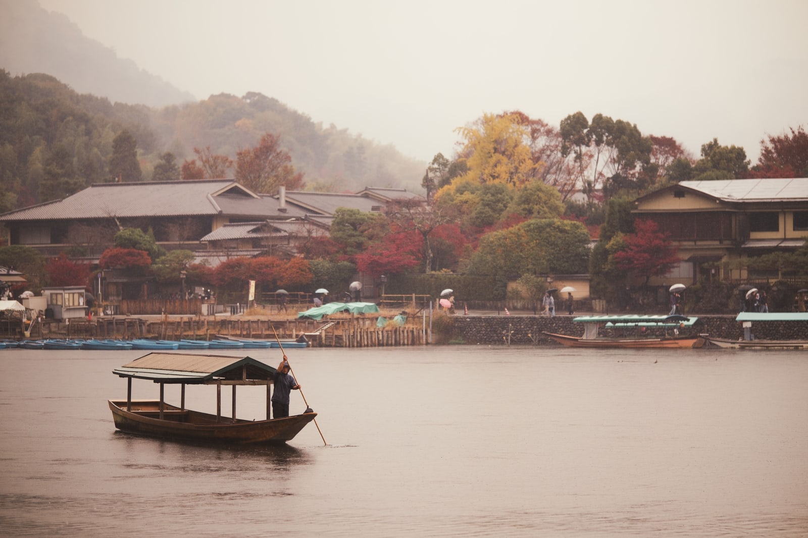 A small roofed boat poled across a wide river in misty rain, tiled houses and autumn trees on the far bank, people with umbrellas on the embankment.