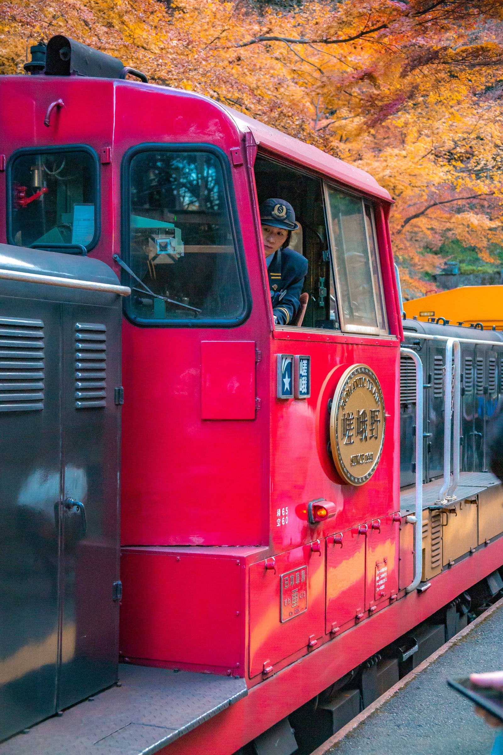 The red diesel locomotive of the Sagano Romantic Train at a platform, its brass headmark reading Romantic Train, the driver leaning from the cab under orange maple leaves.
