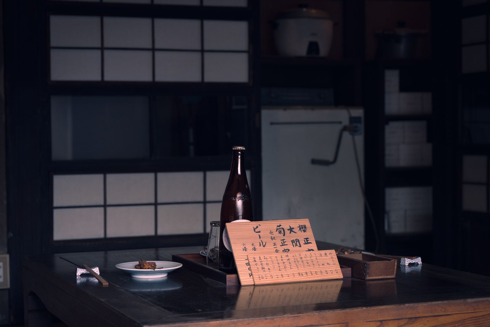 A sake bottle on a dark table beside a hand-written wooden card in a dim room.