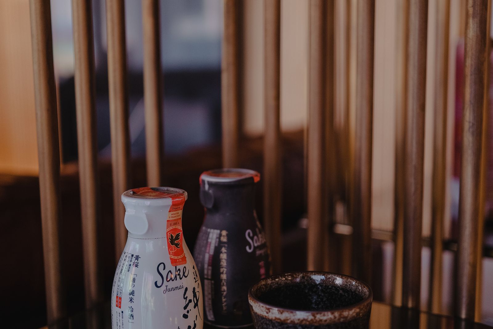 Two sake bottles and a glazed cup on a table, one bottle sealed with a paper excise strip.