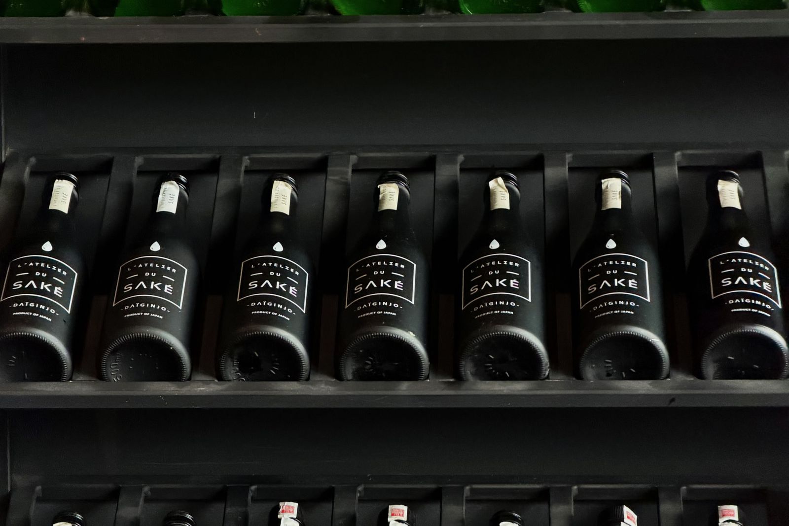 Seven matte black sake bottles on a dark retail shelf, printed in roman type.