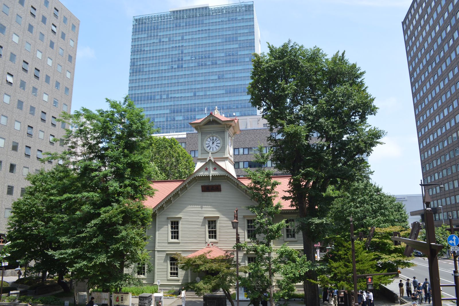 The white wooden Sapporo Clock Tower with its red roof, between summer trees and office towers