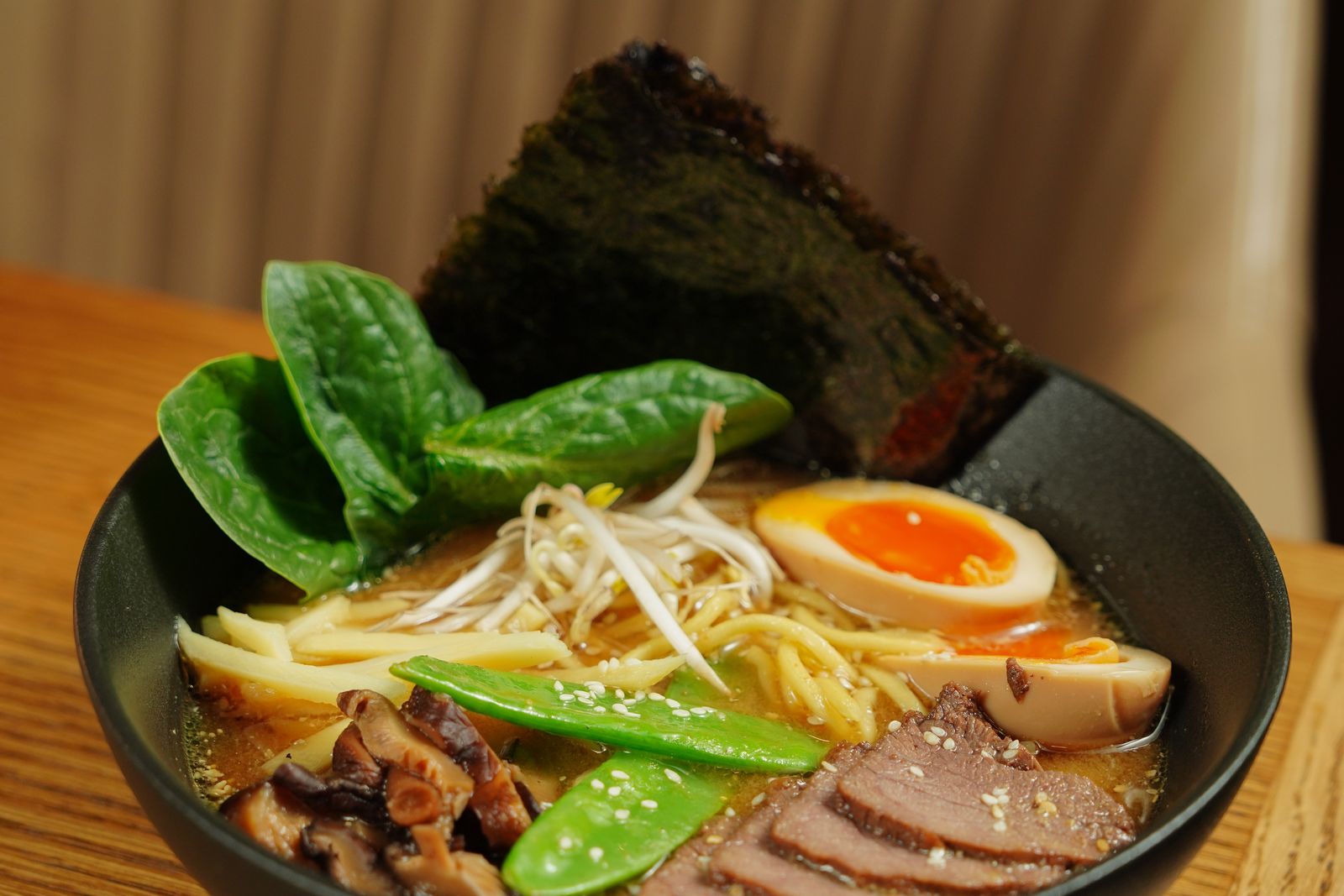 A bowl of ramen in a rich brown broth with bean sprouts, bamboo shoots, greens, a soft egg and nori.