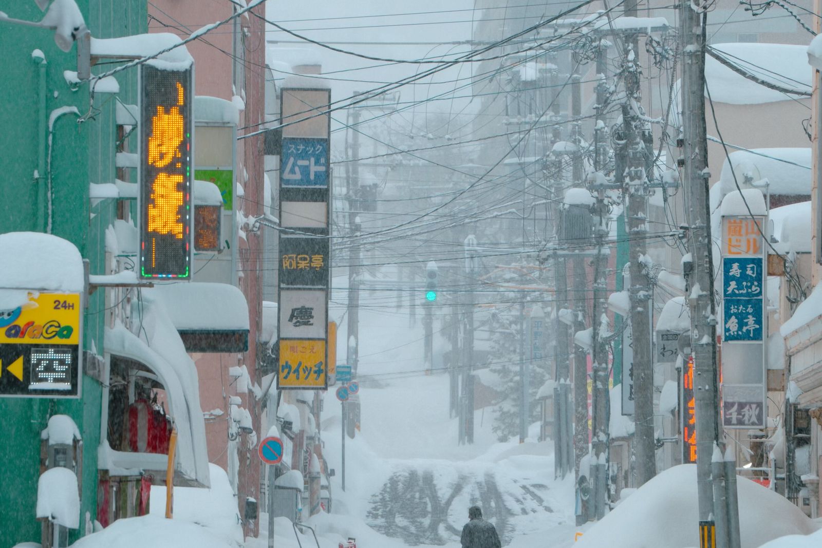 A narrow Hokkaido street under heavy snow, a lone figure pulling a sled up the middle of the road between snow-laden signs and power lines.