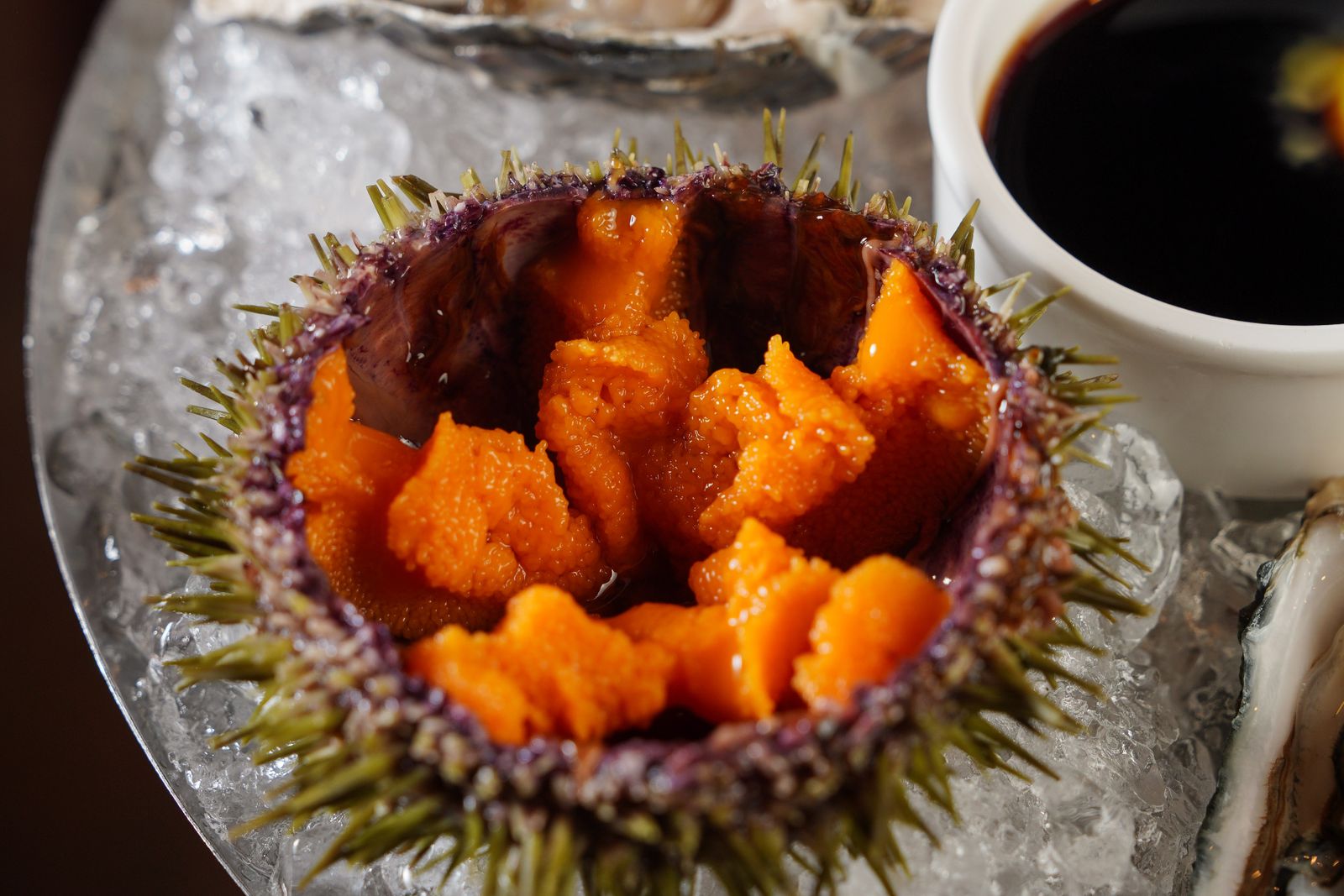 Sea urchin roe in its opened shell on crushed ice, beside an oyster, a lemon wedge and a dish of soy sauce.