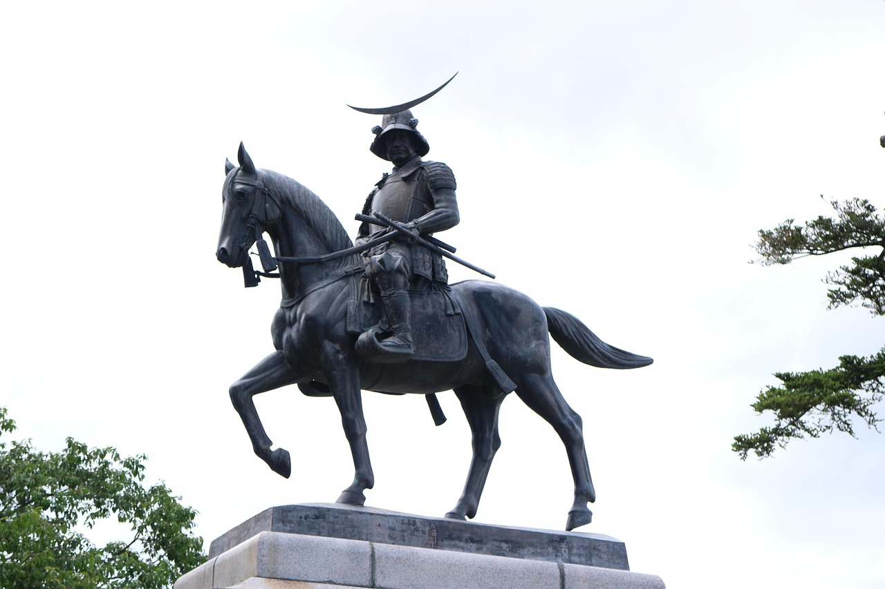 A bronze equestrian statue of a samurai in a crescent-crested helmet on a grey stone base, tree branches at the edges of the frame