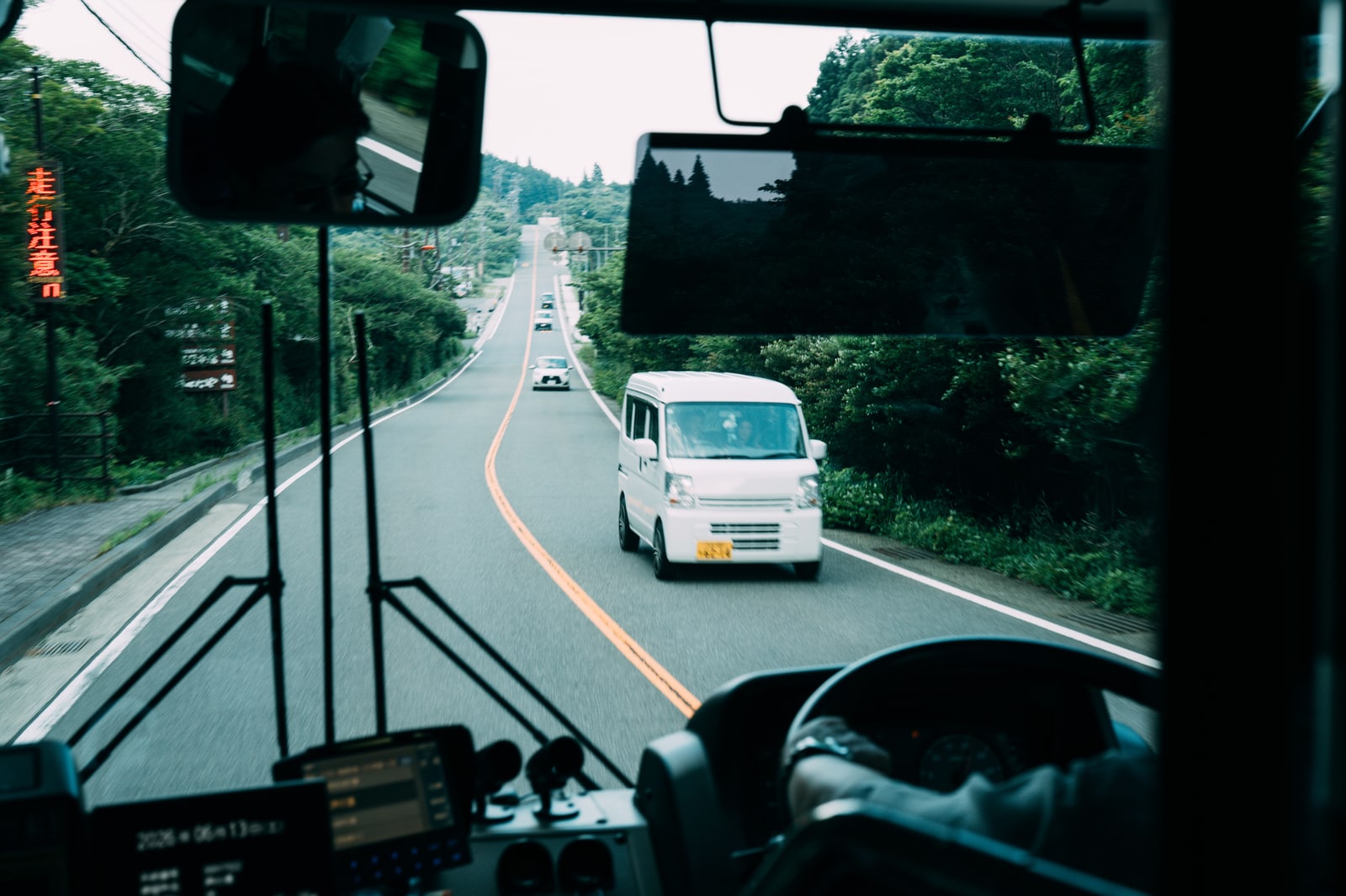 The view through a bus windshield onto a two-lane road through green hills, a small white van ahead.