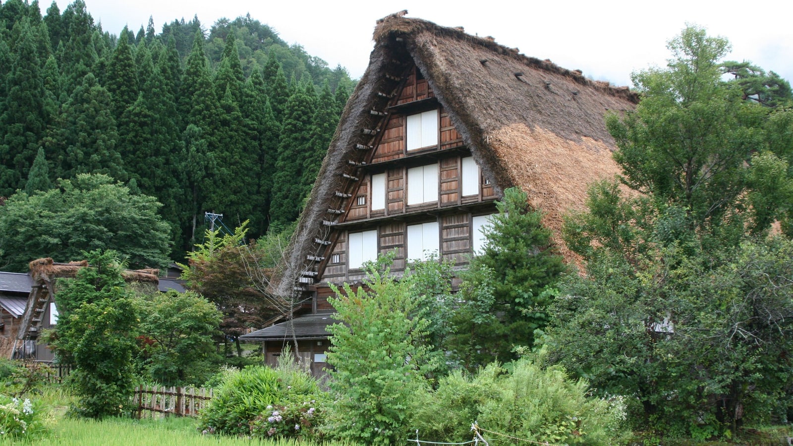 A tall thatched gassho-style house with rows of small windows in its gable, surrounded by trees, in Nanto, Toyama.