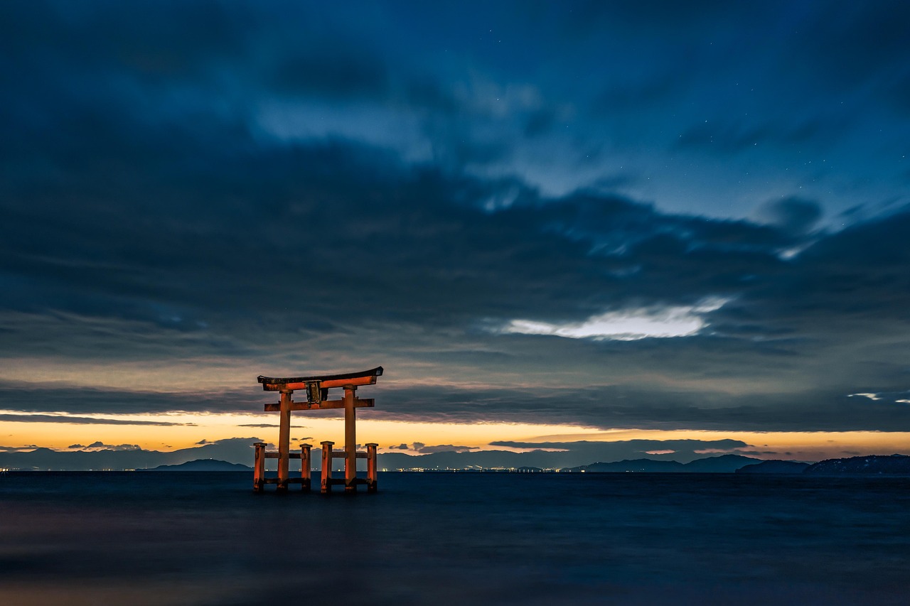 A vermilion torii standing in Lake Biwa before dawn, mountains and shore lights across the water