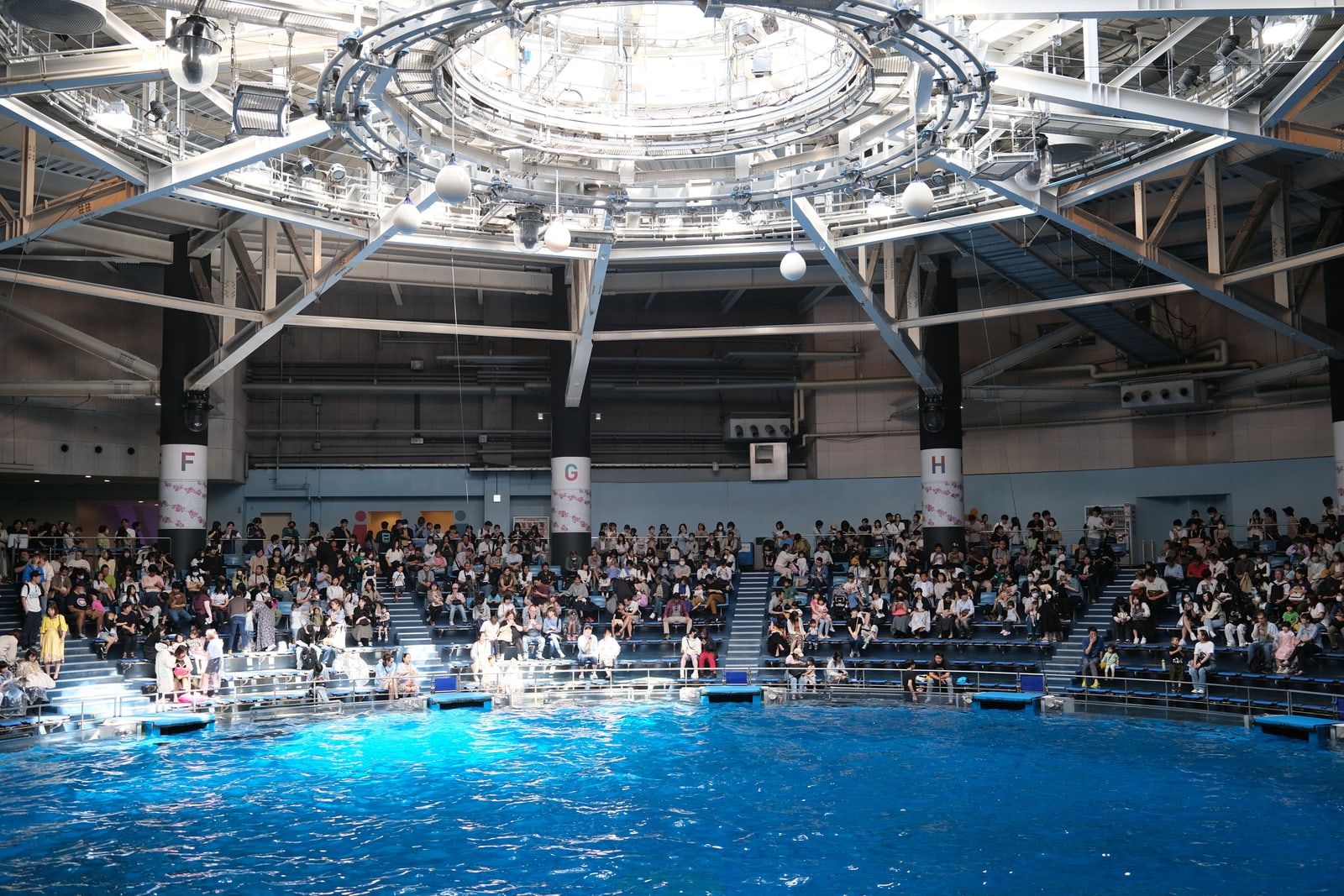 The indoor show pool at Maxell Aqua Park Shinagawa under a steel roof with a circular lighting rig, the tiered seats full of visitors