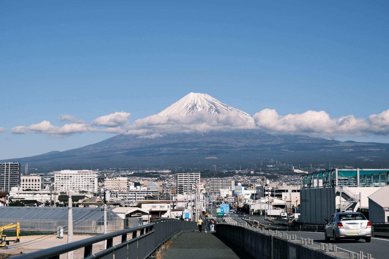 Snow-capped Mt. Fuji above a band of cloud, a low city and a road in the foreground under clear blue sky.