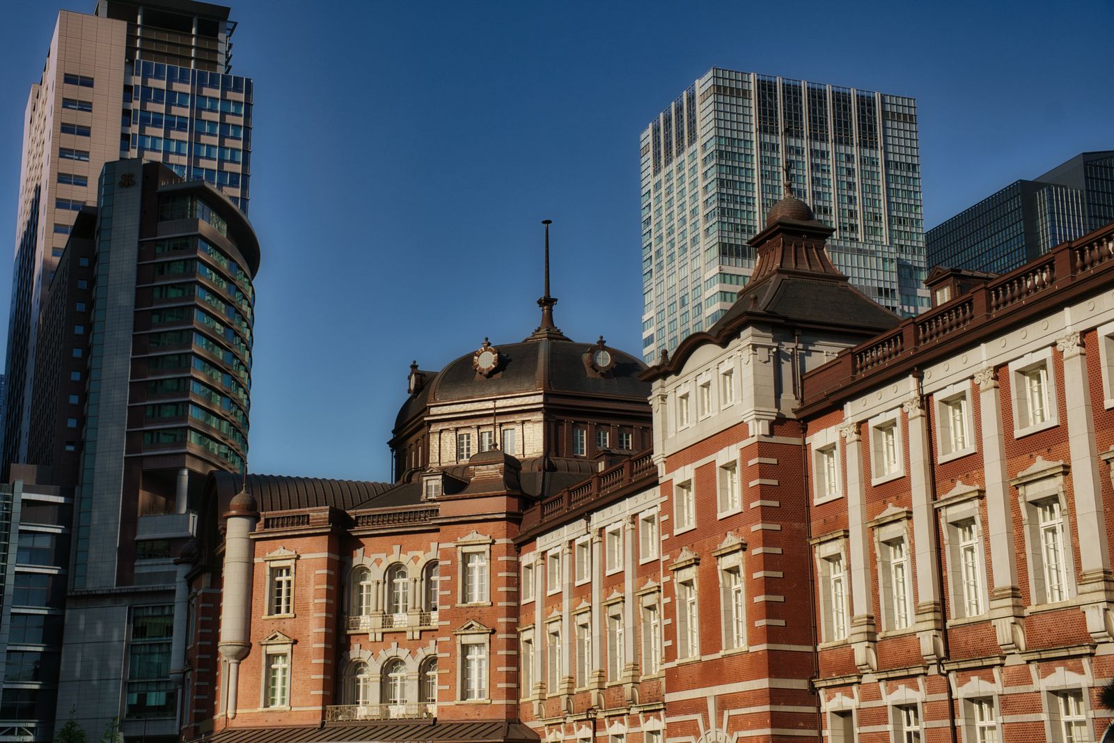 The red-brick facade and domed roof of a historic station building under blue sky, glass office towers rising behind it.