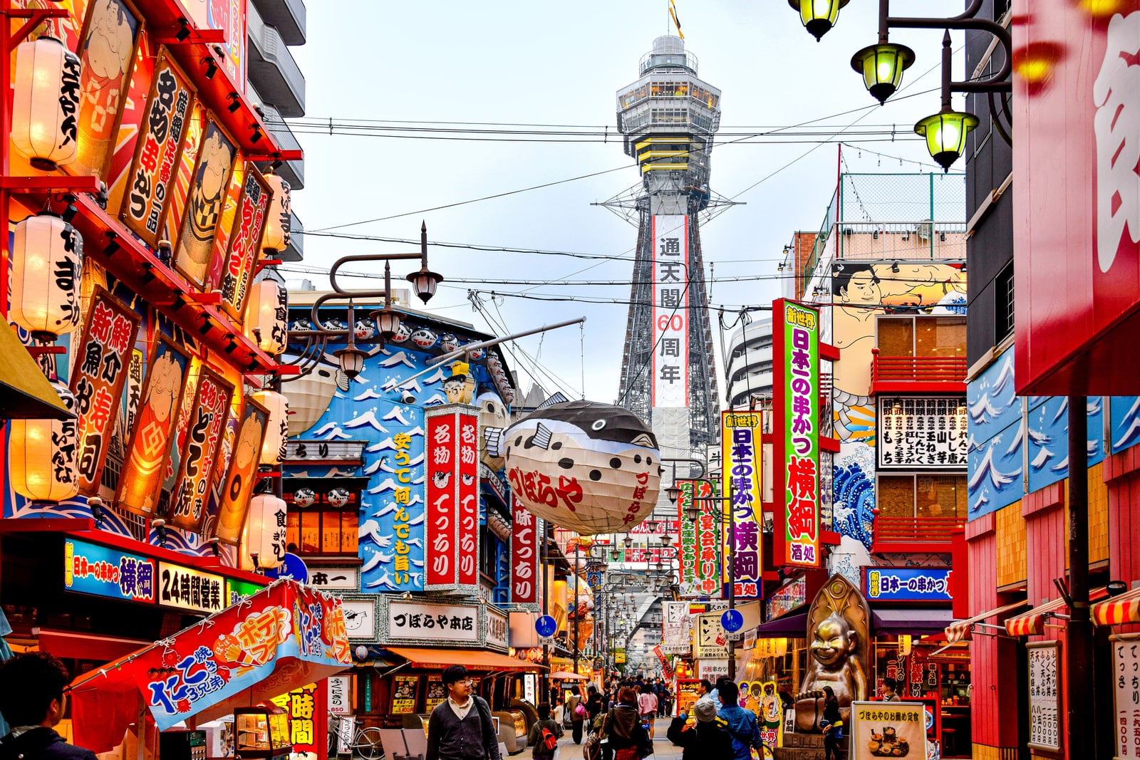 A busy Shinsekai street by day with a giant pufferfish lantern, kushikatsu signs and Tsutenkaku tower in the distance