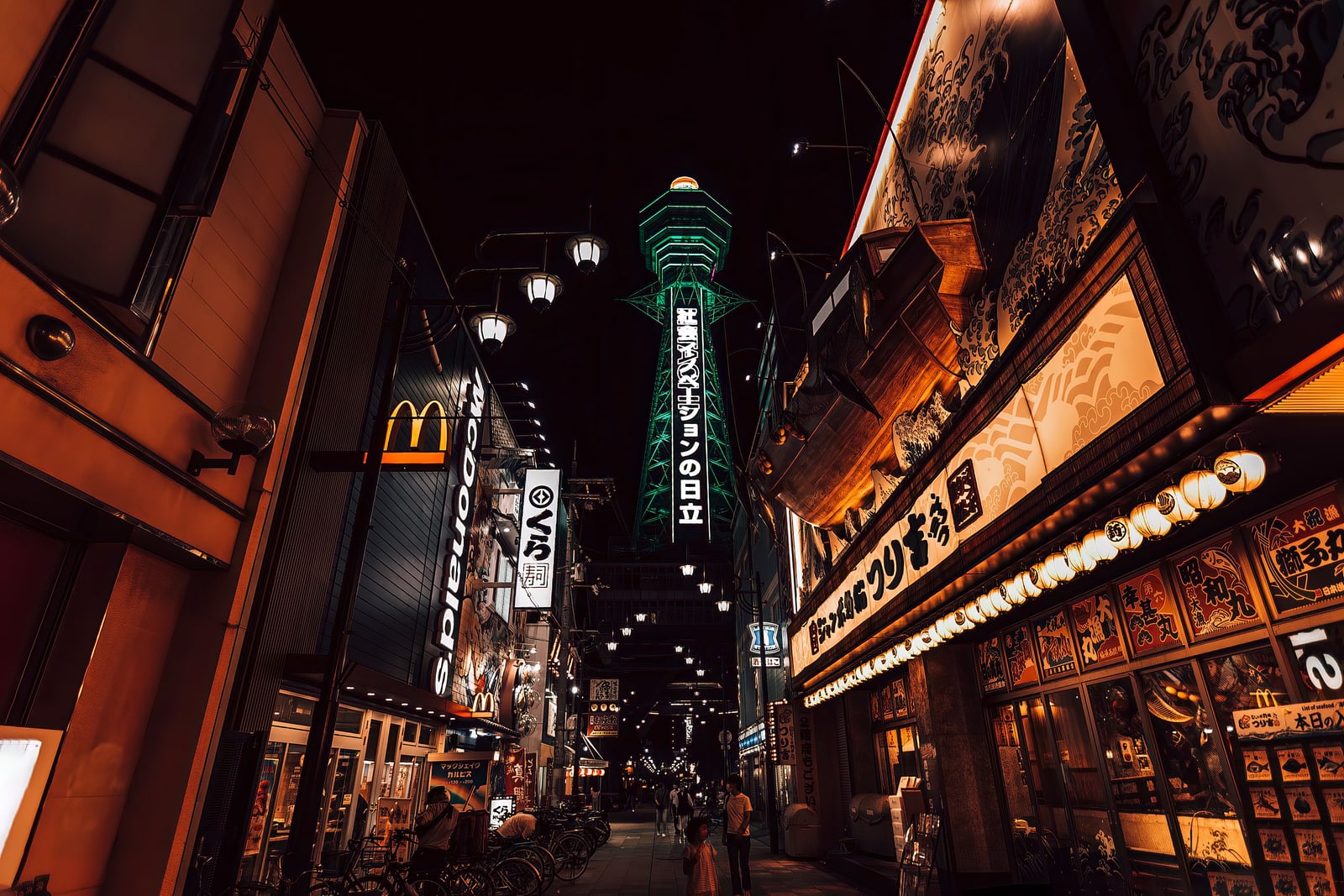 A narrow Shinsekai street at night, Tsutenkaku tower lit green at the far end and lantern-lit restaurant fronts on the right