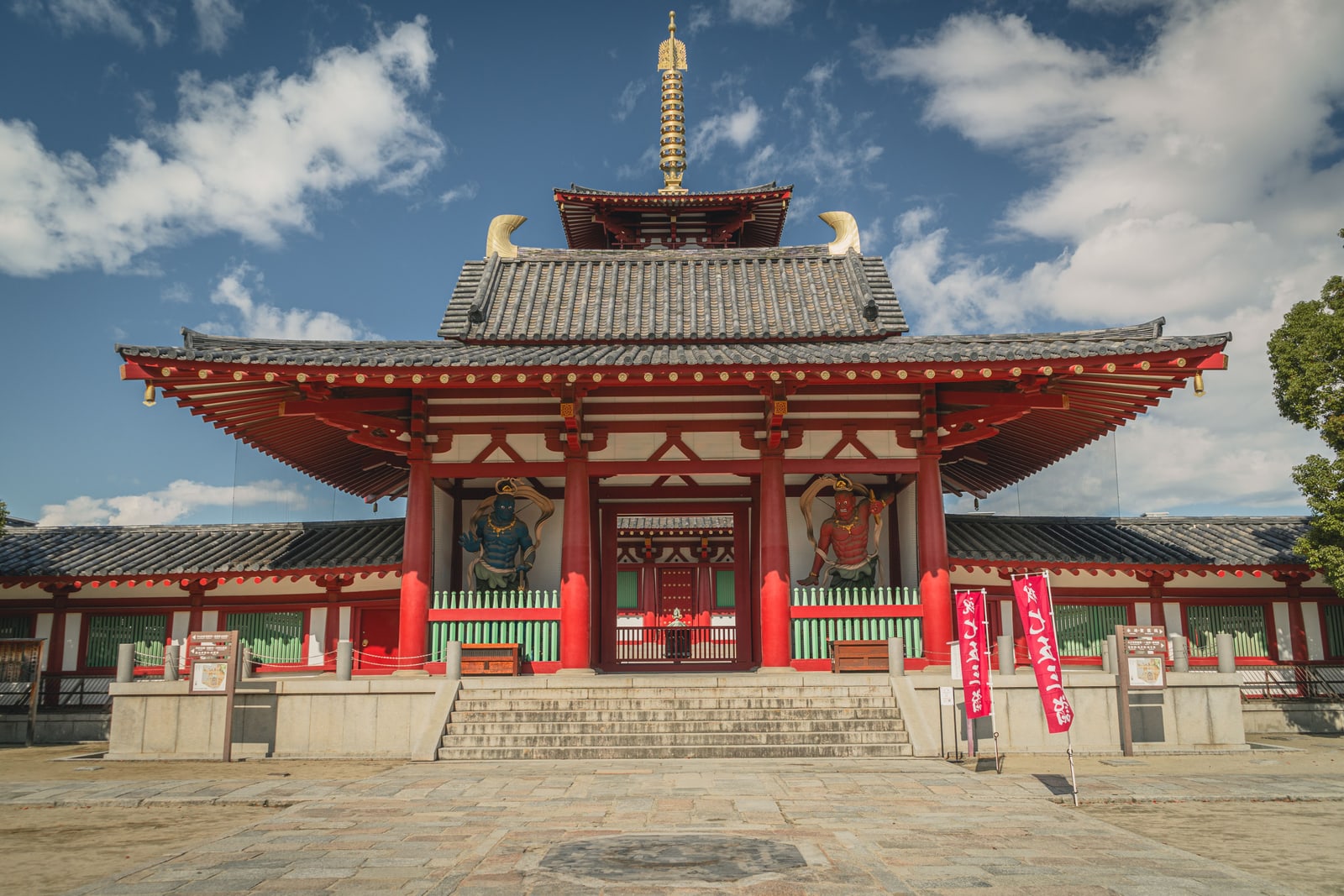 A red-pillared temple gate with a blue and a red guardian statue either side of the opening, stone steps in front and a gold finial rising behind the roof