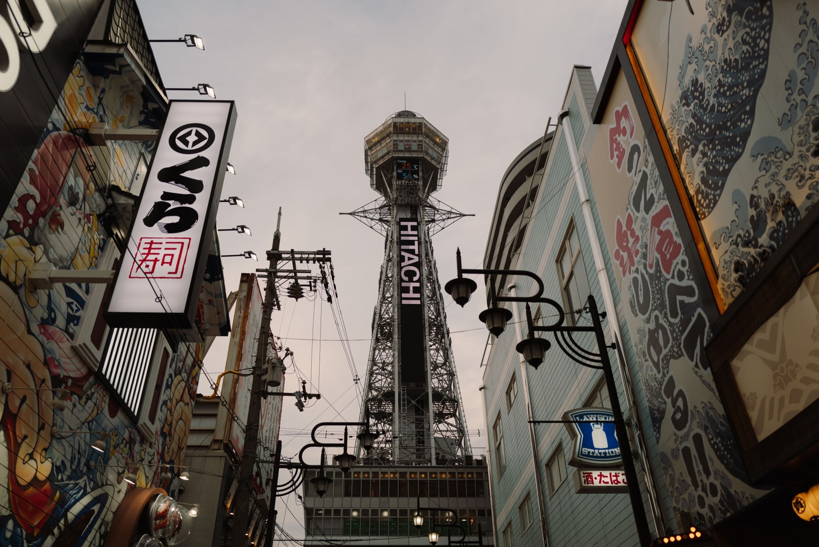 Tsutenkaku tower seen up a narrow Shinsekai street between shop signs and wave-pattern murals at dusk