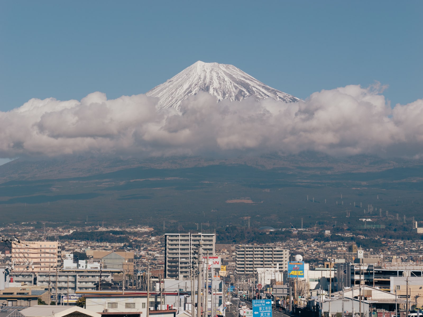 Mt. Fuji rising above a band of cloud over a city on the Shizuoka side, a road sign pointing to Tagonoura Port and Fujinomiya