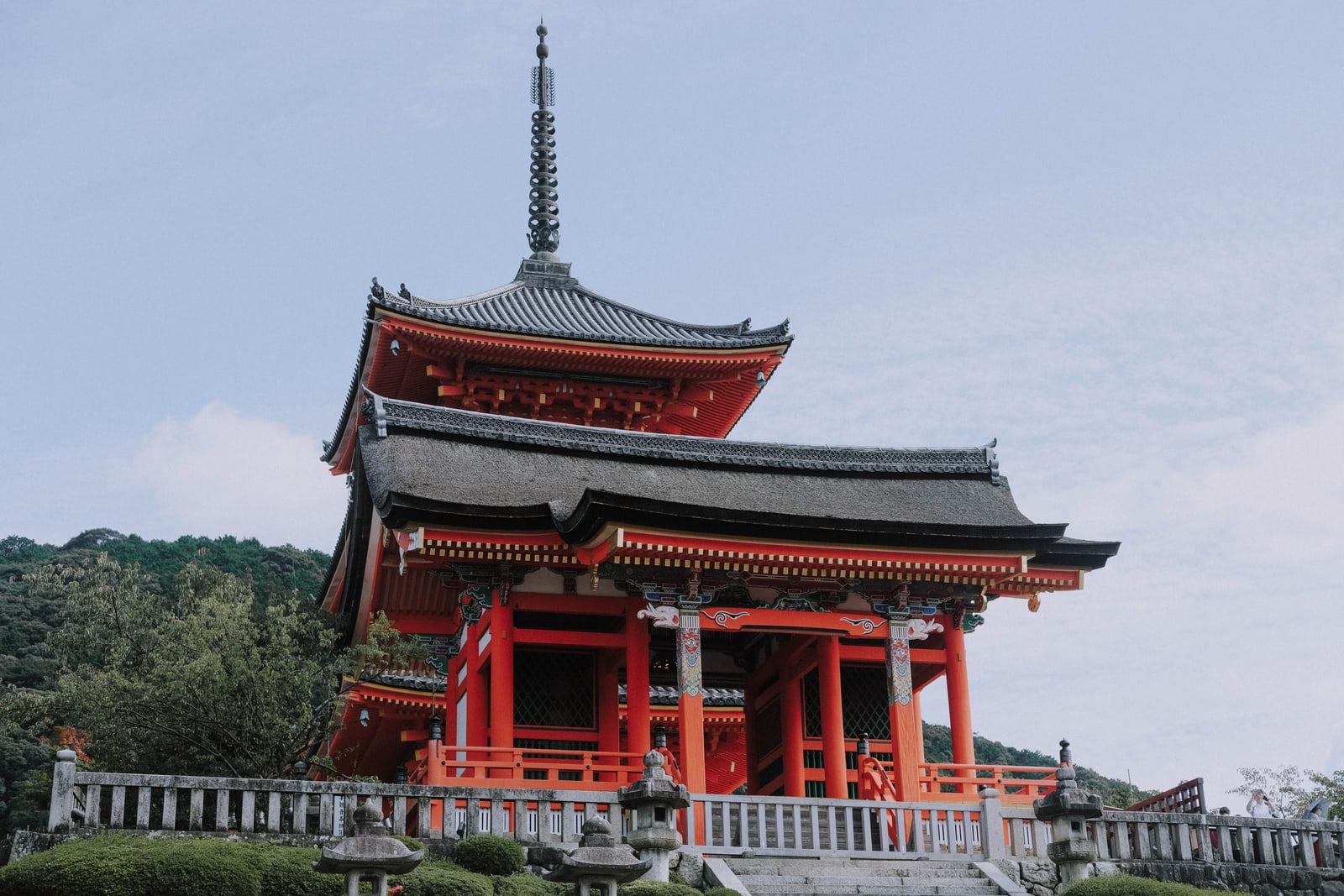 A vermilion-and-white temple pagoda with a tiled roof and bronze spire seen from below against a clear sky, stone lanterns at its base.